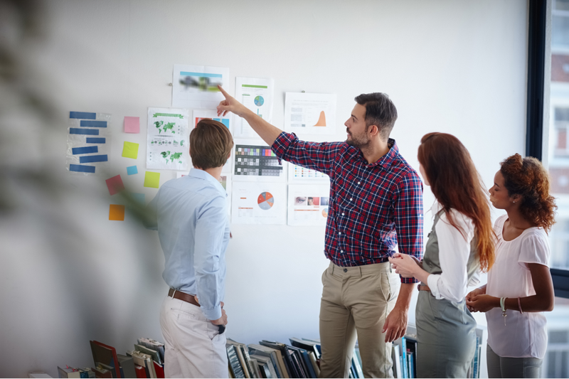 A group of four people are engaged in a discussion while looking at charts and sticky notes on a wall.