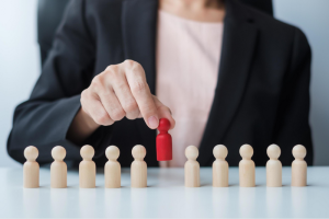 A person in a business suit selects a red wooden figure from a row of beige wooden figures on a table.