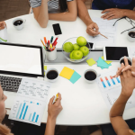 A group of people sitting around a table with laptops, documents, and apples, having a meeting.