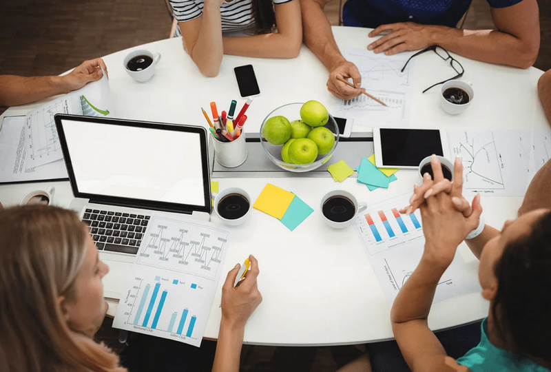 A group of people sitting around a table with laptops, documents, and apples, having a meeting.