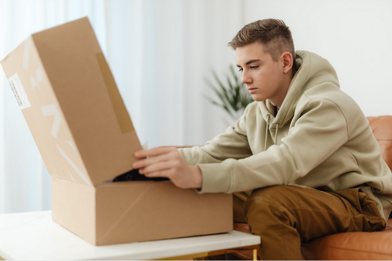 A young man in a hoodie opens a large cardboard box on a table.